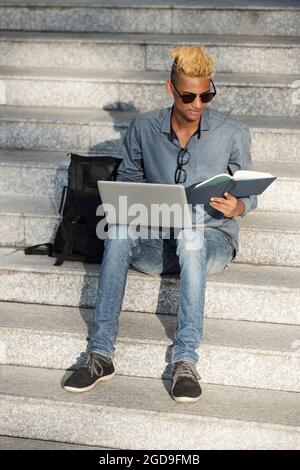 Studente di informatica seduto su passi di edificio universitario, leggere il libro e la codifica sul laptop Foto Stock