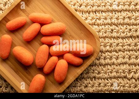 Diverse mini carote naturali arancio luminoso su un piatto di bambù, primo piano, su un tappetino di paglia, vista dall'alto. Foto Stock