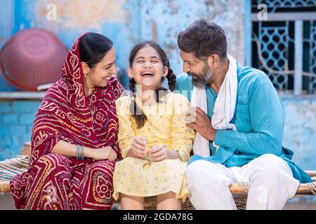 Ritratto di felice famiglia rurale indiana ridendo e avendo un buon tempo insieme mentre si siede fuori a casa del villaggio, allegra piccola figlia godere Foto Stock