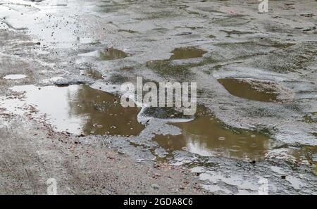 Un buco pieno d'acqua in una strada asfaltata. Una giornata piovosa in una grande città, le auto stanno guidando lungo la vecchia strada. Cattiva strada con asfalto in buche e buche tha Foto Stock