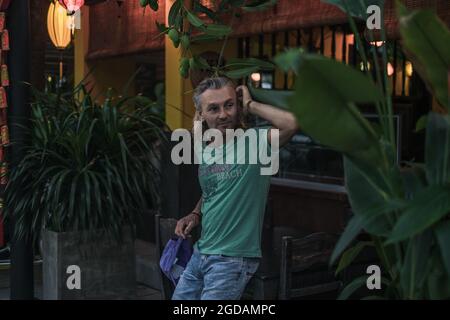 Ritratto di uomo maturo in t-shirt, jeans in piedi e tenendo la testa. Capelli lunghi grigi. Lanterne cinesi. Sera. Alberi tropicali sullo sfondo. Foto Stock