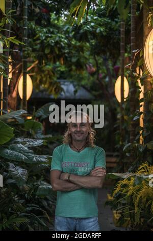 Ritratto di uomo maturo in t-shirt, jeans in piedi sulla strada verde. Capelli lunghi grigi. Lanterne cinesi. Sera. Alberi sullo sfondo. Foto Stock