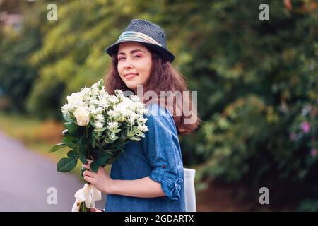 Ritratto di una giovane bella donna in un cappello di paglia che tiene un bouquet di rose bianche. Piante e alberi sullo sfondo. Il concetto dell'8 marzo e Mo Foto Stock