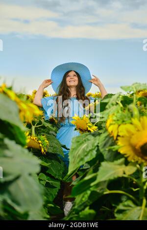 Donna in abito blu cielo tiene i bordi del cappello di paglia e guardando in su tra girasoli Foto Stock