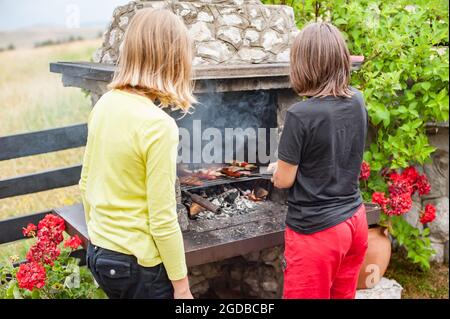 Carne alla griglia per bambini. Ragazzo e ragazza che fanno barbecue sulla griglia sulla natura. Campeggio per famiglie e barbecue Foto Stock