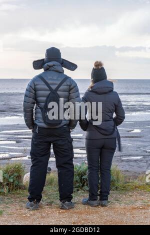 coppia vestita di caldo abbigliamento invernale in piedi su una spiaggia che guarda al mare sulla costa del norfolk, coppia che guarda all'orizzonte, abiti invernali, coppie Foto Stock