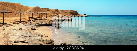 Costa sabbiosa e rocciosa con ombrelloni da spiaggia di palme. Spiaggia desolata resort sulla costa del Mar Rosso. Sharm el Sheikh, Sinai, Egitto, Asia in estate Foto Stock