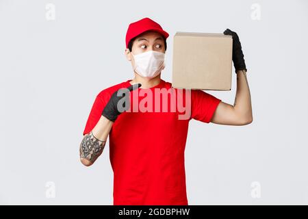 Concetto di consegna e di trasporto durante la pandemia di coronavirus. Intrigato tipo di consegna in uniforme, servizio di corriere portare scatola al cliente, indicando e. Foto Stock