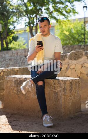 Un giovane che usa un telefono cellulare. È seduto su una panca di pietra. È caucasico con pelle marrone. Indossa una T-shirt gialla, pantaloni neri e. Foto Stock