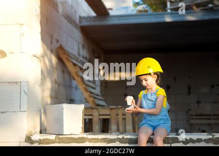 La ragazza piccola in un hardhat giallo sta giocando il costruttore sul luogo di costruzione della sua casa futura. Attesa di muoversi, scegliere una professione, bambini Foto Stock