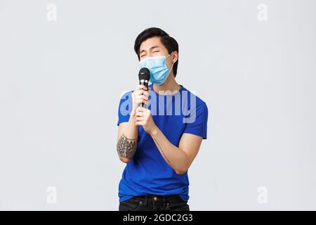 Stile di vita Covid-19, emozioni della gente e tempo libero sul concetto di quarantena. Carefree giovane ragazzo coreano divertente, uomo asiatico in maschera medica che tiene il microfono Foto Stock