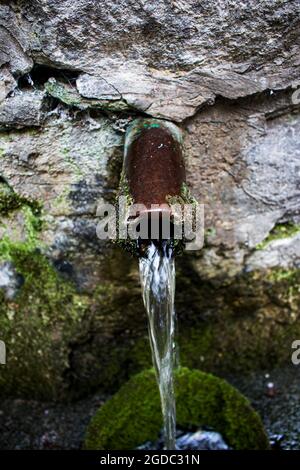 L'acqua proveniente da una sorgente forestale scorre attraverso un tubo metallico rivestito di pietre. Fonte naturale di acqua pulita. Foto Stock