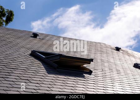 Un ritratto di un lucernario aperto in un tetto d'ardesia in una giornata di sole con un cielo blu con nuvole bianche Foto Stock