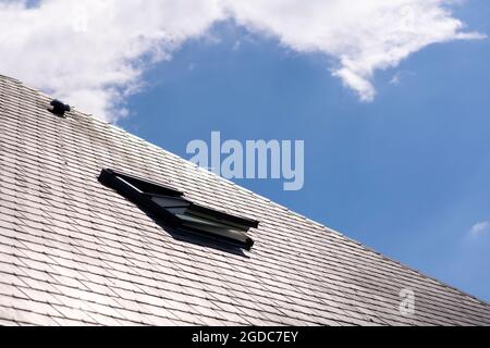 Un ritratto di un lucernario aperto per la ventilazione in un tetto di ardesia in una giornata di sole con un cielo blu con nuvole bianche Foto Stock
