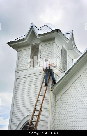 Giovane uomo saldi su una scala appoggiata contro una chiesa nel nord Arkansas. Egli è nel processo di aiutare a reroof una vecchia chiesa. Foto Stock