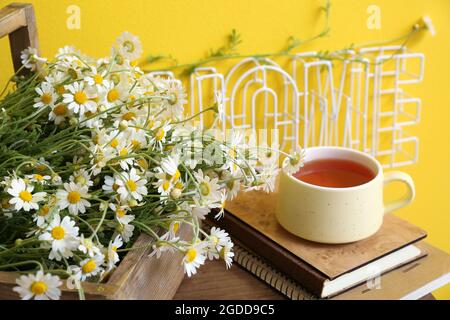 Scatola con camomiles, libri e tazza di tè sul tavolo vicino parete colorata Foto Stock