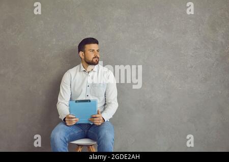 Giovane uomo seduto sulla sedia, tenendo il resume e guardando via la parete grigia dello spazio della copia Foto Stock