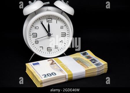 un pacco di banconote da 200 euro si trova di fronte a un orologio bianco con le mani impostate a 5 minuti a 12 Foto Stock