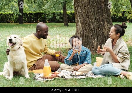 Famiglia africana seduta sull'erba e mangiando mentre si passa il tempo di picnic insieme al loro cane Foto Stock
