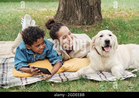 Madre felice sdraiata sull'erba con suo figlio usando il telefono cellulare e giocando con il loro cane nel parco Foto Stock