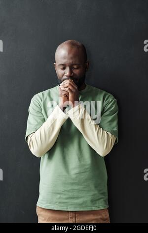 Ritratto di giovane africano che prega con gli occhi chiusi mentre si trova in piedi sullo sfondo nero Foto Stock