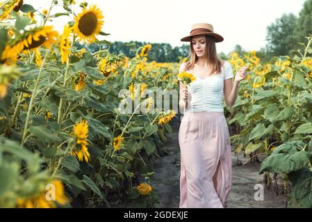 bella ragazza dolce in un cappello di paglia che cammina su un campo di girasoli sorridenti Foto Stock