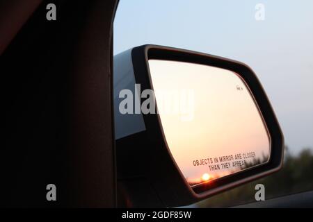 Rückspiegel eines Autos mit Abendsonne und dem Hinweis: Gli oggetti nello specchio sono più vicini di quelli che appaiono. Foto Stock