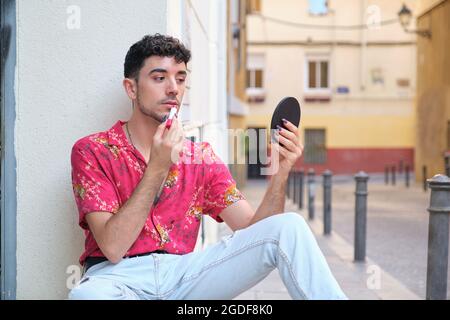 Giovane uomo caucasico con lunghe false unghie applicare rossetto rosso per strada. Foto Stock