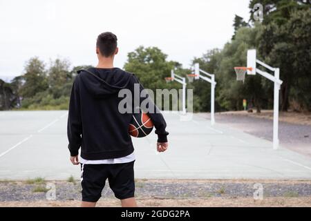Un adolescente di sesso maschile del giocatore di basket guardando fuori oltre il campo di pallacanestro, vista posteriore Foto Stock