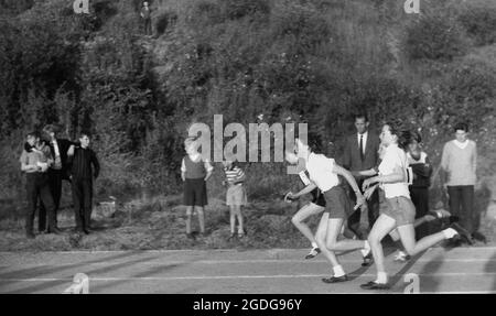 1964, storico, fuori dal comune in una giornata di sport tra le scuole della contea, le ragazze che gareggiano nella corsa di staffetta, i corridori in arrivo passano il testimone al prossimo corridore in attesa in pista, Exeter, Devon, Inghilterra, Regno Unito. Foto Stock