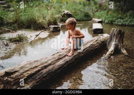 Ragazzo giovane seduto sul log in acqua in estate guardando premuroso Foto Stock