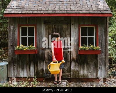 Ragazzo annaffiando fiori in finestre scatole di capannone con grande annaffiatura CAN. Foto Stock