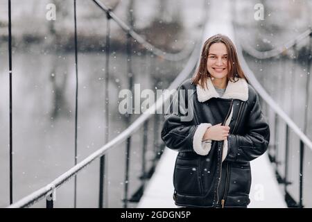 Una donna cammina sul fiume su un ponte sospeso in inverno. Giovane ragazza in abiti caldi si trova su un ponte di legno in una fredda giornata innevata. Foto Stock