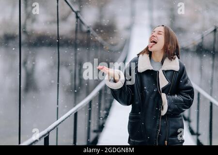 Una donna cammina e cattura fiocchi di neve con la lingua sopra il fiume su un ponte sospeso in giorno d'inverno. Giovane ragazza in abiti caldi si alza su un legnino Foto Stock