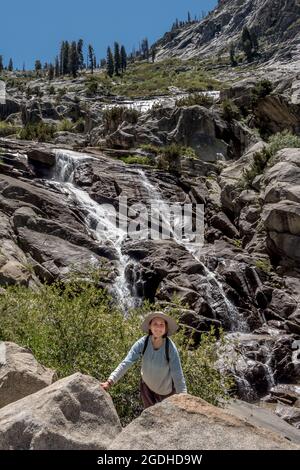 Una ragazza che sorride per una foto con la parte inferiore della cascata Tokopah Falls mentre si camminano nel Sequoia National Park. Foto Stock