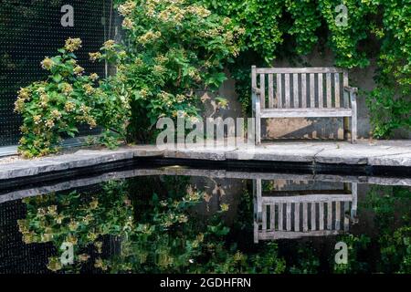 Una panca di legno si trova sotto il verde fogliame vicino ad un laghetto su un percorso concreto Foto Stock