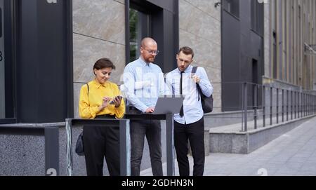 Attraenti uomini d'affari caucasici che parlano usando il laptop e discutono il progetto mentre la donna che parla o chiacchiera usando il telefono delle cellule all'aperto nel centro. Architettura o design Foto Stock