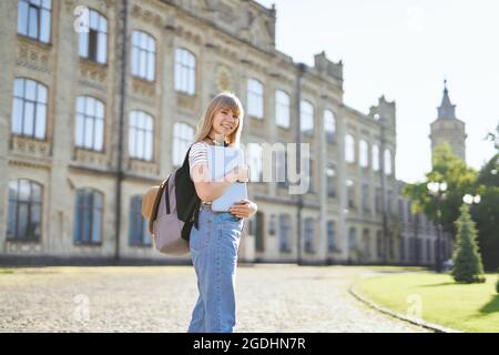 Adorabile giovane studentessa bionda universitaria o studentessa di scuola superiore con zaino e computer portatile indossando jeans in piedi nel campus universitario. Concetto educativo o di studio. Immagine di alta qualità Foto Stock