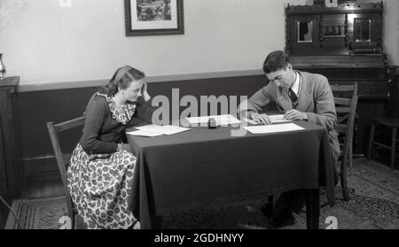 anni '50, storico, all'interno di una sala da pranzo dell'epoca, una coppia seduta su sedie di legno alla fine di un tavolino, coperto da una tovaglia. Utilizzando una penna stilografica e con una bottiglia d'inchiostro sul tavolo, l'uomo iis scrivere su fogli di carta, mentre la giovane donna sta leggendo il manoscritto manoscritto manoscritto Foto Stock