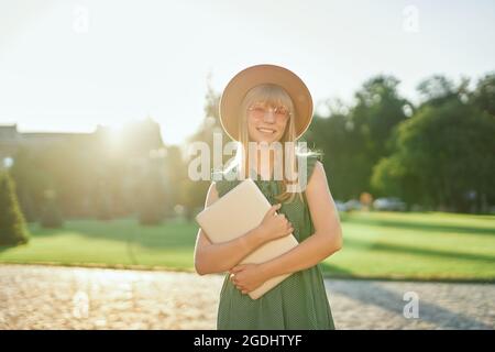 Adorabile giovane studentessa bionda universitaria o universitaria con un computer portatile che indossa un abito verde e un cappello nel campus universitario. Felice scuola femminile o studente, concetto educativo. Immagine di alta qualità Foto Stock