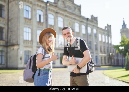 Due studenti allegri, maschi e femmine, con zaini che sollevano i pollici al campus universitario tenendo un laptop. Adorabile coppia di studenti di successo sorridendo mostrando i pollici fino segno alla fotocamera Foto Stock