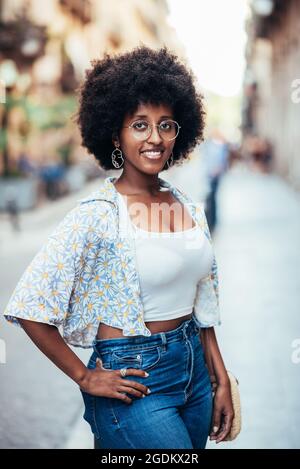 Giovane donna africana che si trova per le strade di Barcellona e guarda la macchina fotografica, sta posando con una mano sull'anca e sorridendo Foto Stock