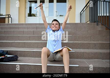 Felice studente elementare alzando le braccia con la matita in mano, si sente felicità e soddisfazione dopo il lavoro di realizzazione. Concetto di ritorno a scuola Foto Stock