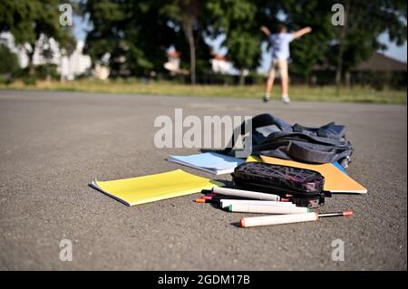 Gli approvvigionamenti scolastici sparsi, il contenitore a matita, i pennarelli, i libri da lavoro che cadono da uno zaino aperto, giacciono sull'asfalto del cortile contro lo zaino Foto Stock