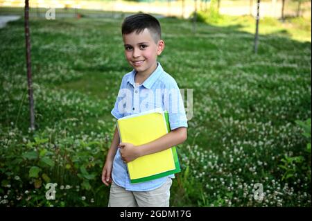 Felice studente elementare che torna a scuola. Adorabile scolaro che tiene un libro da lavoro e sorride alla macchina fotografica in piedi su sfondo verde erba. Foto Stock