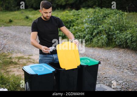 Uomo che lancia il sacchetto di plastica nel cestino di riciclaggio sul cortile posteriore. Colori diversi sui contenitori in plastica. Verde, blu e giallo Foto Stock