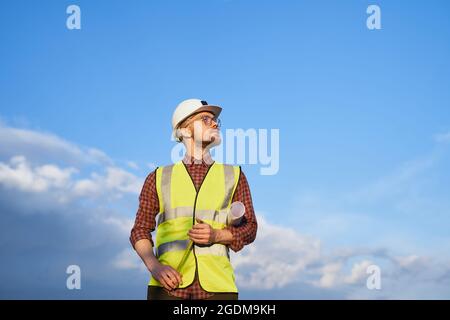 Vista ad angolo basso dell'ispettore maschio bearded, del supervisore o dell'appaltatore dell'edificio nel casco e del gilet riflettente di sicurezza con nastro-misura su un cantiere. Giornata di sole con cielo blu. Immagine di alta qualità Foto Stock