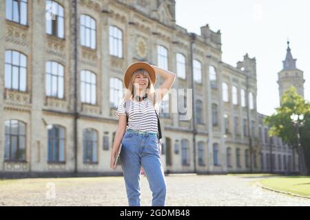 Carino felice università femminile o ritratto studentesco dell'università. Studiare all'estero o concetto educativo. Allegro giovane bionda con zaino e laptop indossando jeans e cappello nel campus. Immagine di alta qualità Foto Stock