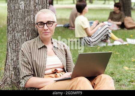 Ritratto di giovane donna d'affari in occhiali guardando la fotocamera mentre si lavora su un computer portatile nel parco Foto Stock
