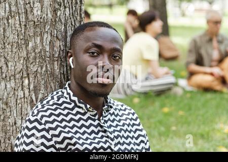 Ritratto di un giovane africano in cuffie wireless che ascolta la musica e guarda la fotocamera mentre sei seduto nel parco Foto Stock